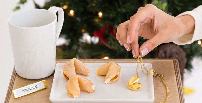 Fortune Cookies on a plate and someone is placing down a Fortune Cookie Locket next to them. Behind the table tis a Christmas tree and the table has a mug and fortune Frame that reads
